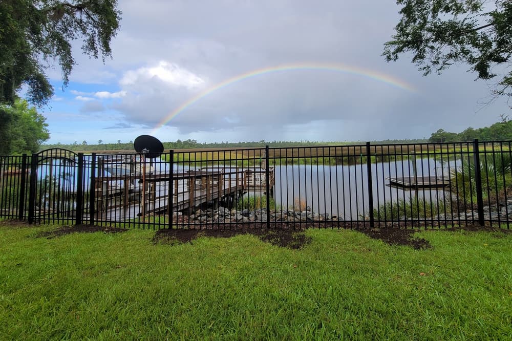Black aluminum fence with an arched gate along a tidal creek, a rainbow over the marsh behind it