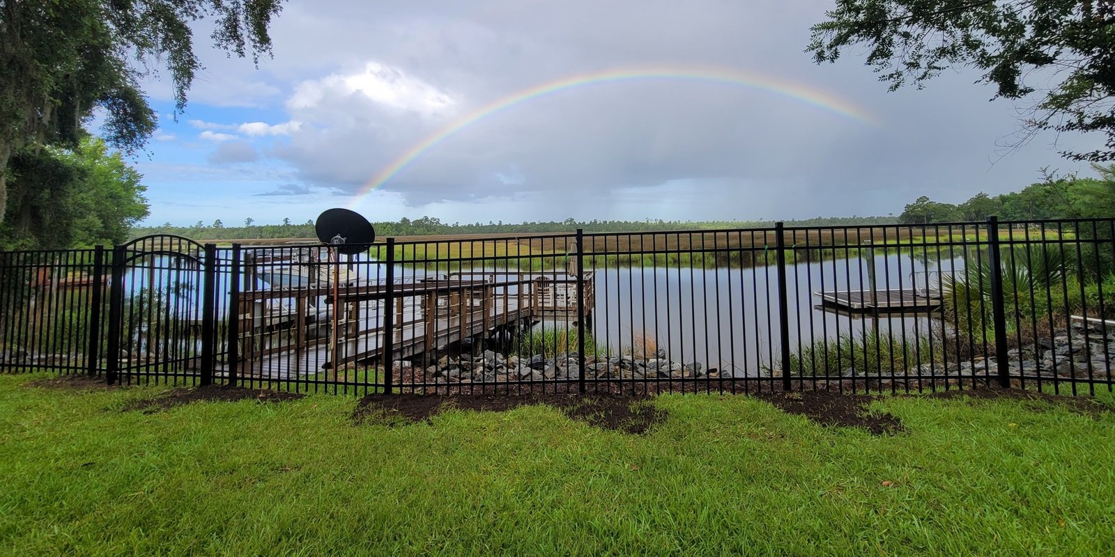 Black aluminum fence along a mowed lawn with a rainbow arching over tidal marsh and a weathered dock