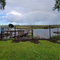 Black aluminum fence along a mowed lawn with a rainbow arching over tidal marsh and a weathered dock