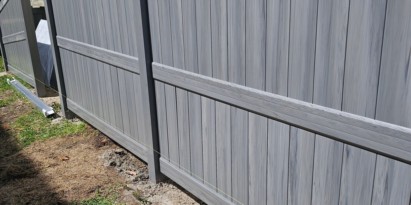 Gray woodgrain vinyl privacy fence beside a stucco building with palm fronds on sandy ground