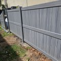 Gray woodgrain vinyl privacy fence beside a stucco building with palm fronds on sandy ground
