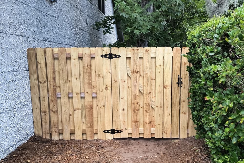 Wood shadowbox gate with heavy black strap hinges hung between a house wall and a hedge