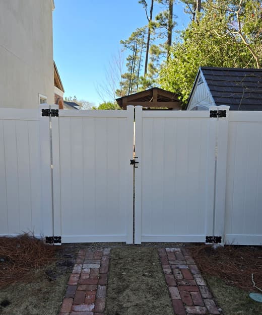 White vinyl privacy fence with a double gate over a brick path between a house and a garden building