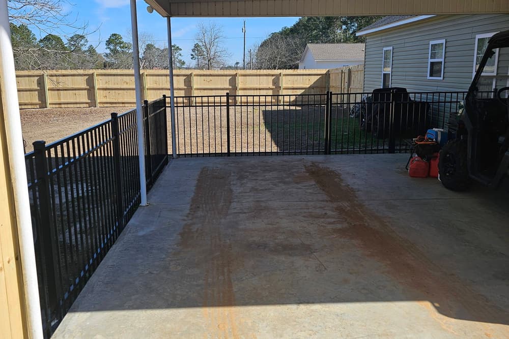 Black aluminum fence and gate enclosing a covered carport slab beside a house
