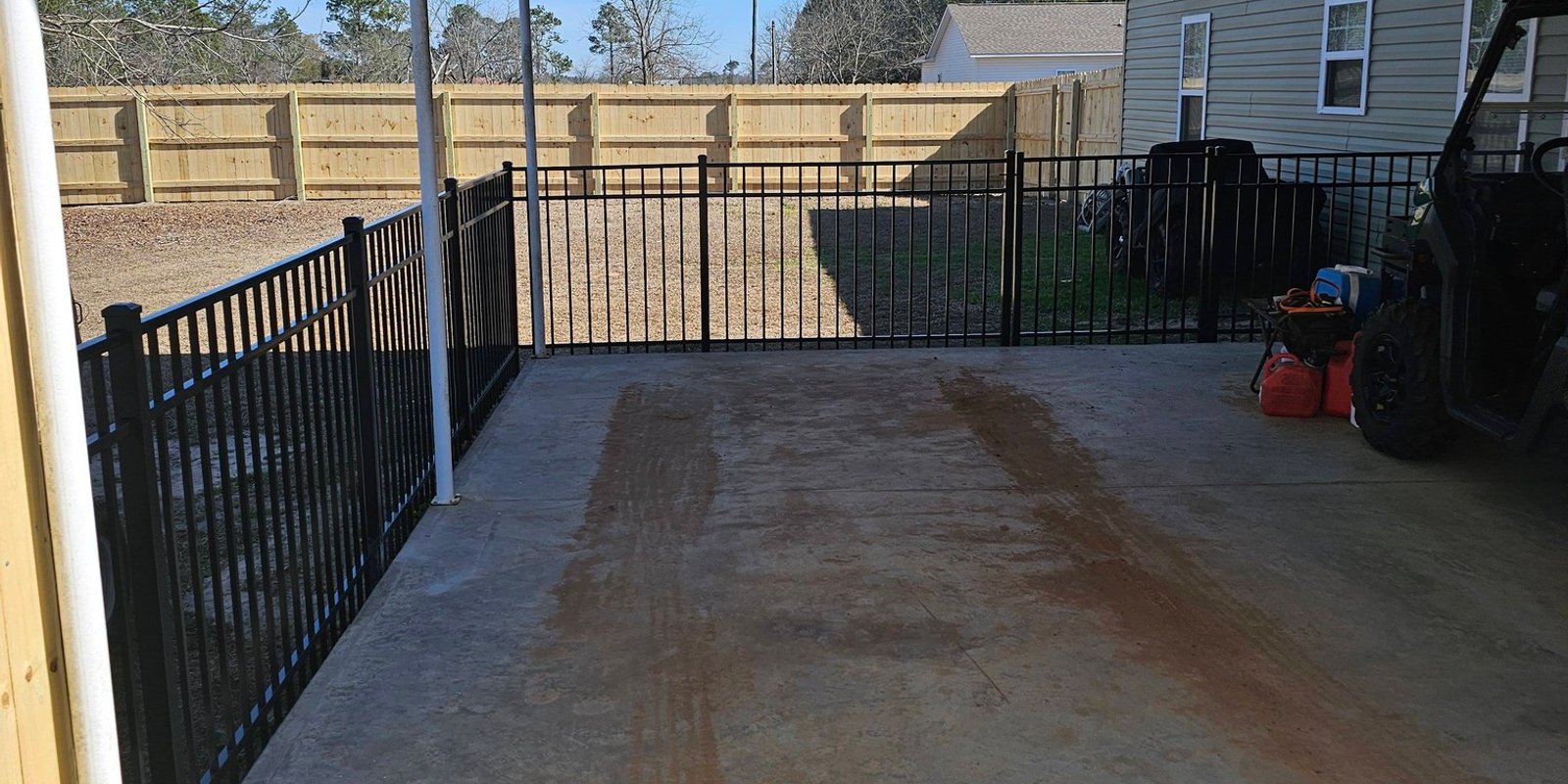 Black aluminum fence and gate enclosing a concrete carport slab with a wood privacy fence beyond