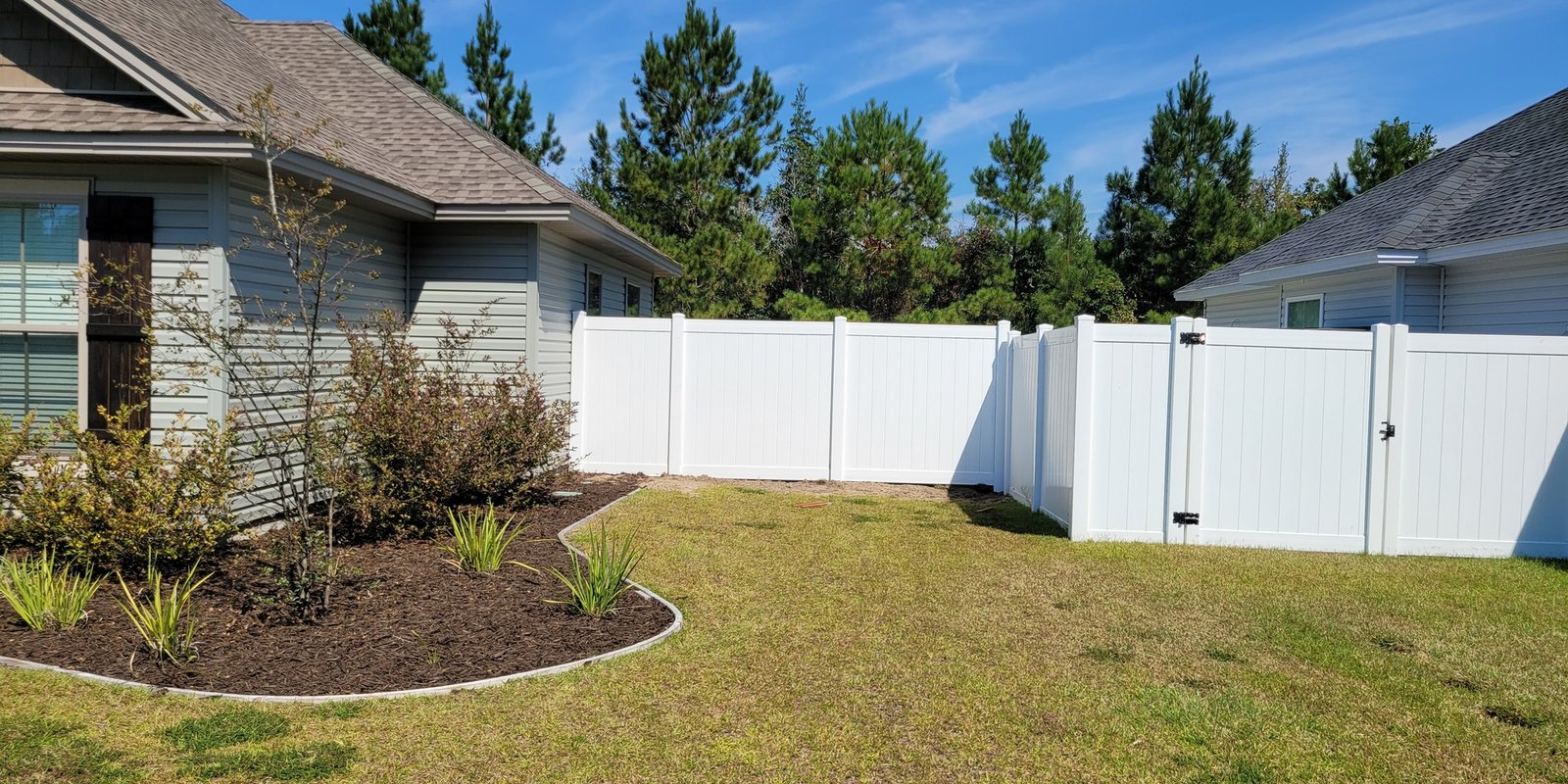 White vinyl privacy fence and gate enclosing a new backyard with a mulched bed and tall pines