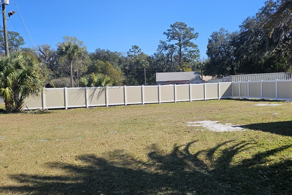 Tan and white vinyl privacy fence enclosing a large open lot with palms and pines behind