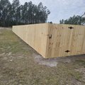 New dog-ear wood privacy fence with a closed gate running across an open lot beside planted pines