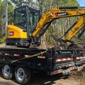 Yellow mini excavator strapped to a black dump trailer on a sandy track through tall pine woods